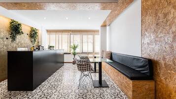 Un área de recepción moderna con un mostrador negro, baldosas de piso con diseño, una zona de estar con sofá y sillones, y un ventanal con persianas de madera.