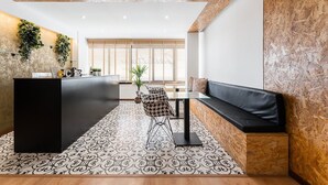 Ein moderner Empfangsbereich mit einem schwarzen Empfangstresen, farbenfrohen Bodenfliesen, einer Sitzecke mit Sofa und Sesseln sowie einem großen Fenster mit Holzjalousien.