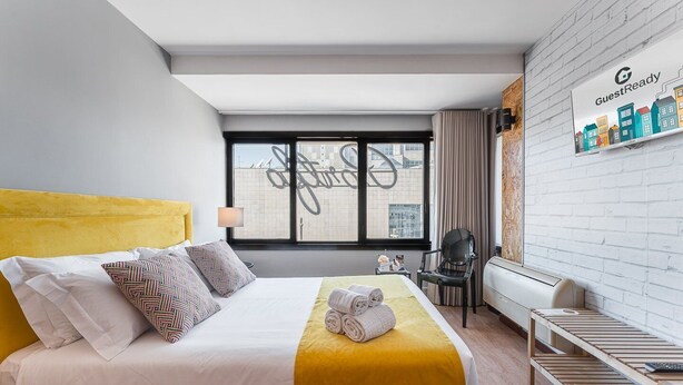 Ein modernes Hotelzimmer mit einem gelben Bett, einem großen Fenster mit Blick auf die Stadt, einem schwarzen Sessel und einer Wand-Klimaanlage.