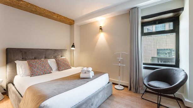 A modern hotel room with a large bed, a bedside lamp, a chair, and a view of the city through a window. - GuestReady Economy- Portfolio Guesthouse Room 407 (Porto)