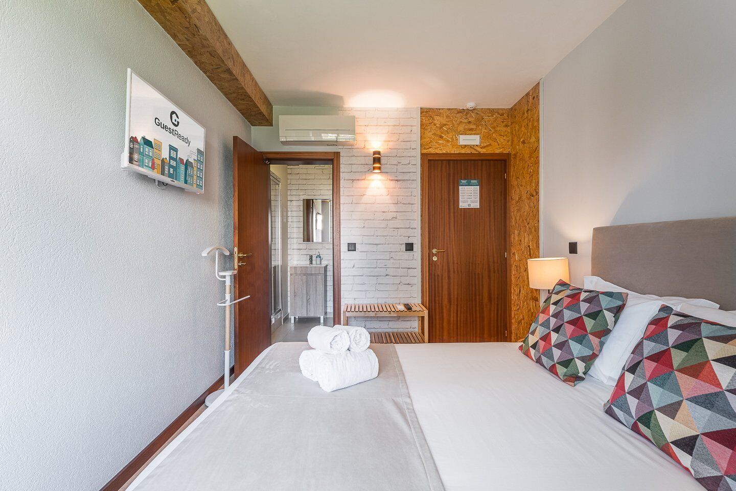 Quarto de hotel com cama, um banquinho, banheiro com espelho e um aviso na parede.