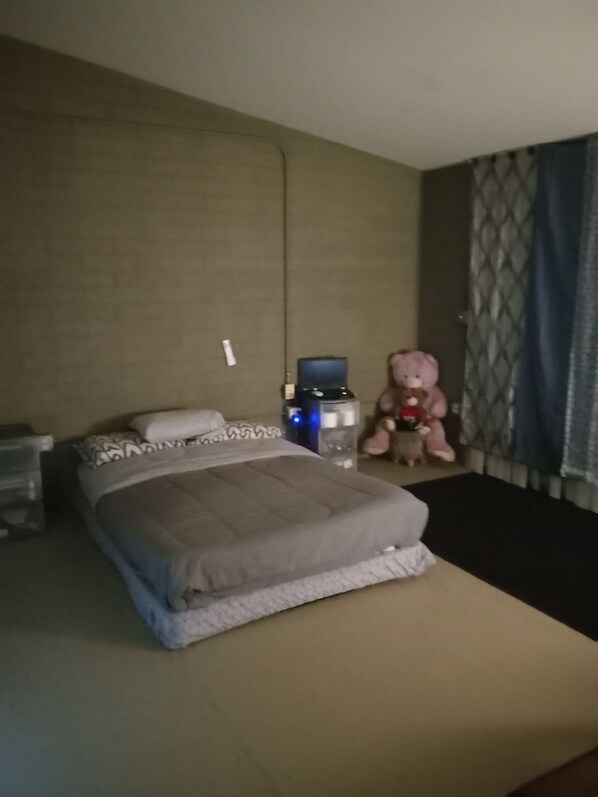 A hotel room with a bed, a small table with a teddy bear, a water bottle, and a power outlet on the wall.