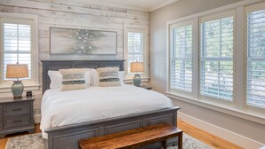 A bedroom with a large bed, bedside tables, a bench, and large windows with blinds.