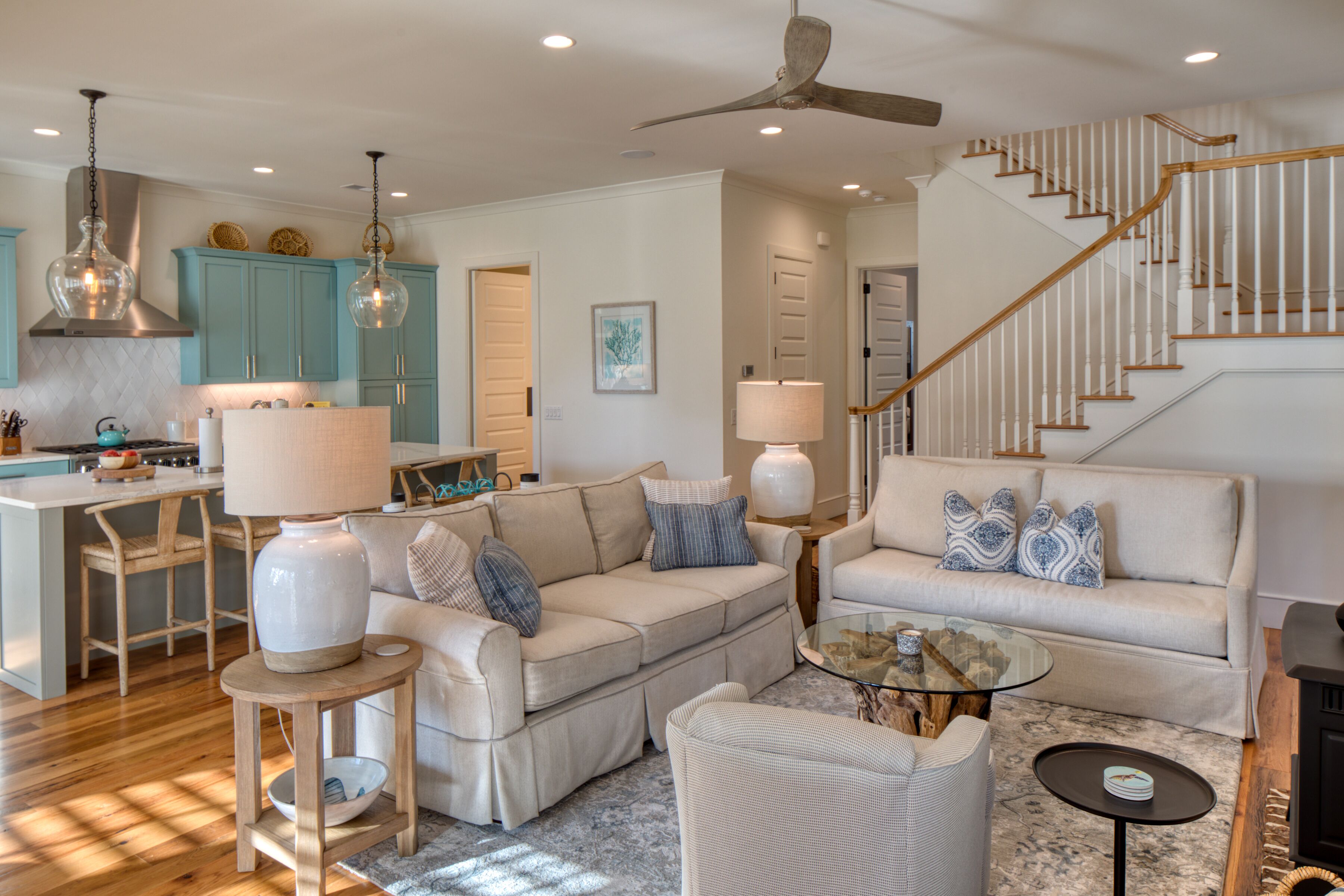 A spacious living area with a sofa, armchairs, a coffee table, and a fireplace. There is a kitchen area with teal cabinets and a dining table. A staircase with wooden railings is visible.