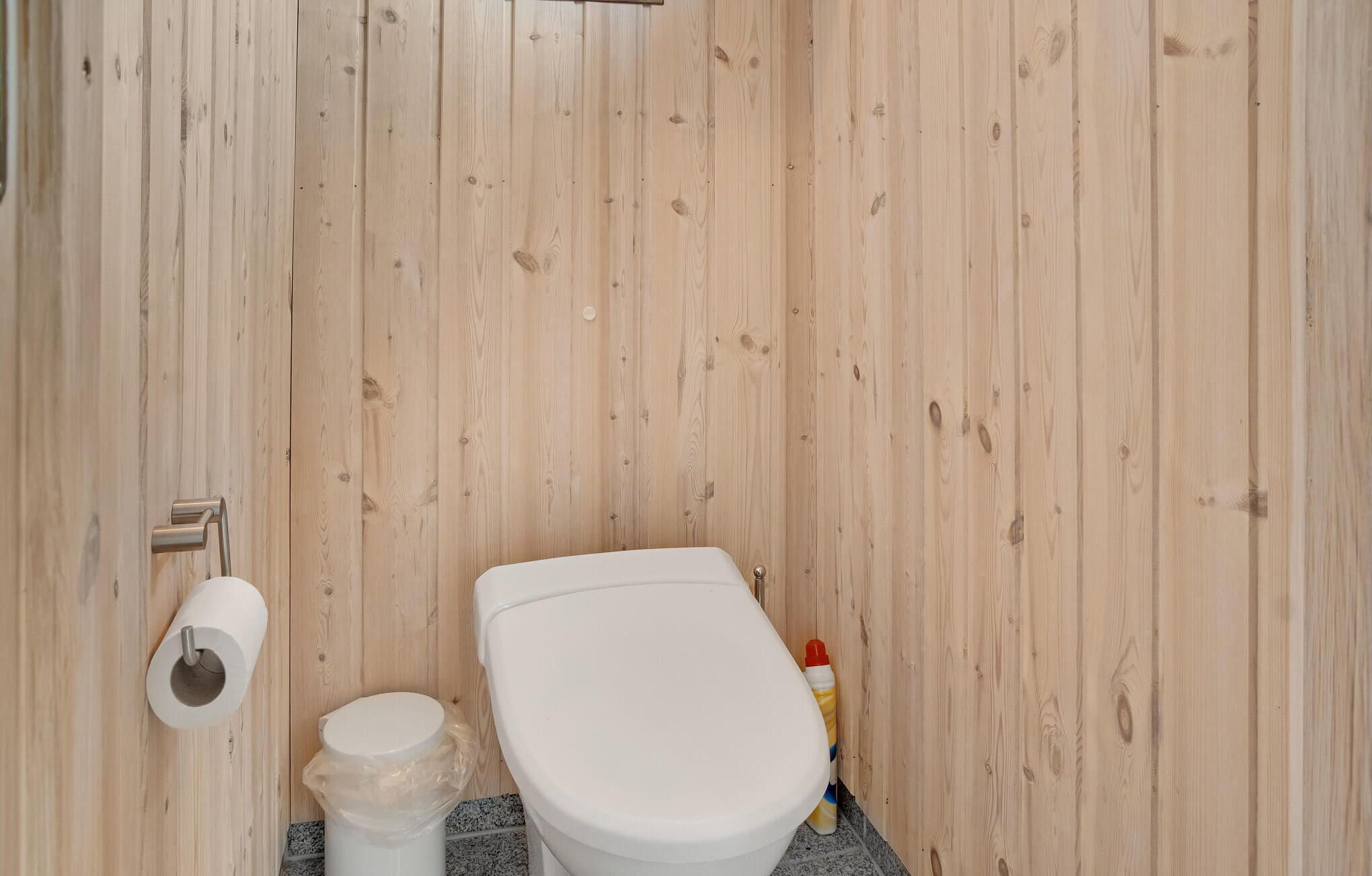 Un baño pequeño con paneles de madera, un inodoro, un dispensador de papel higiénico y una botella de plástico.