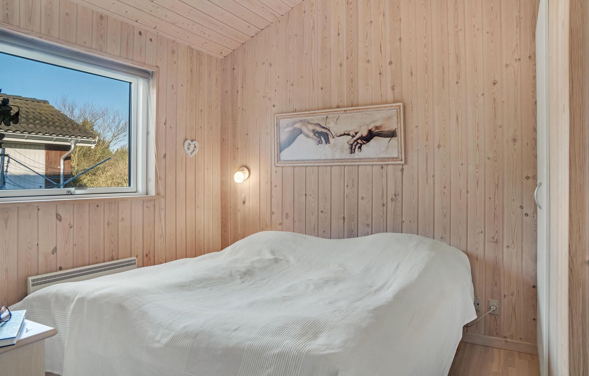 Un dormitorio con paredes de madera, una cama con cubierta blanca, una ventana y un cuadro de manos extendidas.