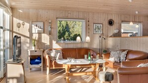 A cozy cabin interior with wooden walls, a large window with a view of greenery, a wooden dining table, and leather seating. - Stunning home in Rødby with sauna (Rødby)