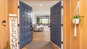 A hallway with two large doors, a welcome sign, a macrame plant hanger, and a view into a living area with a sofa and a coffee table. - Amazing Escape with Game Room, Pool & Hot Tub (Escondido)