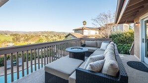 A balcony with a wicker sofa, a round table, and a view of a pool and distant landscape. - Amazing Escape with Game Room, Pool & Hot Tub (Escondido)
