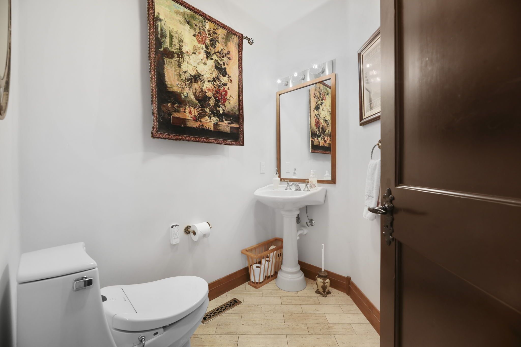 A bathroom with a toilet, sink, and mirror. There is a painting on the wall and a wooden basket near the sink.