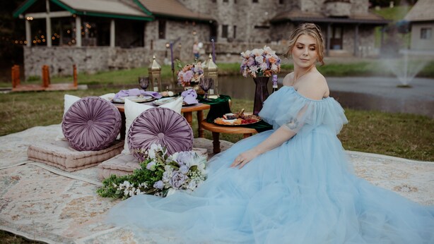 En kvinna i en ljusblå klänning sitter på en picknickfilt vid ett bord som är dukat med blommor, ljus och mat.