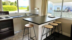 A modern kitchen with a central island, bar stools, a stove, and a view of the outside. - Pohara's Seaside Chalet - Cosy Beach Stay (Pōhara)