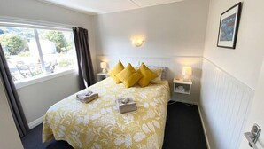 A hotel room with a bed, two bedside lamps, a window with curtains, and a framed picture on the wall. - Pohara's Seaside Chalet - Cosy Beach Stay (Pōhara)