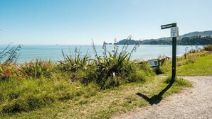 A beach access sign with a shared pathway sign, surrounded by grass and plants, leading to a beach with calm water and distant hills. - Pohara's Seaside Chalet - Cosy Beach Stay (Pōhara)