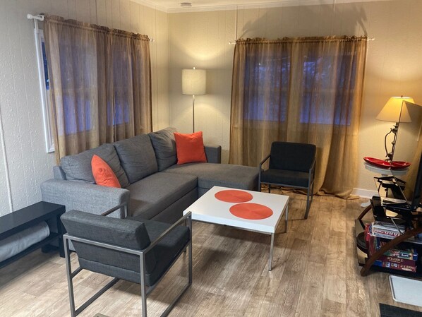 Un salón con un sofá seccional gris, dos sillones, una mesita blanca con círculos rojos, una mesita auxiliar con una lámpara y una estantería con juegos de mesa.