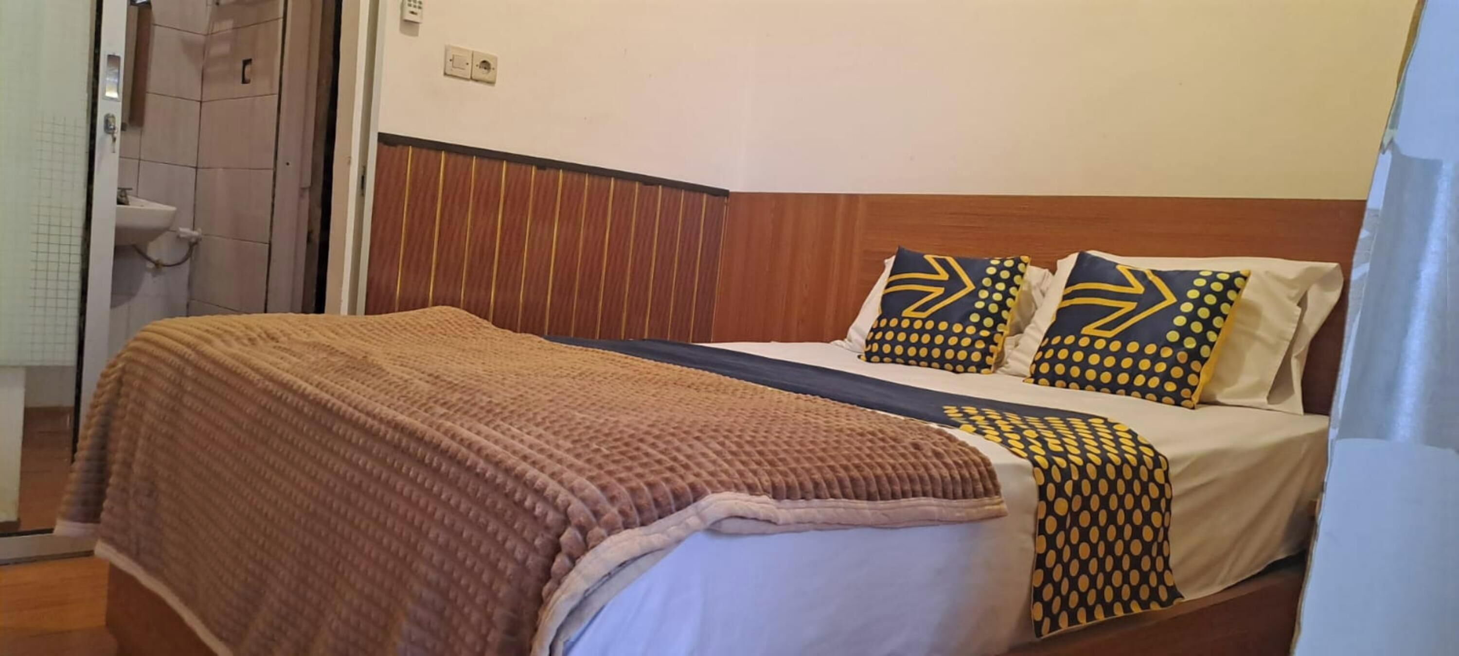 A single bed with a brown blanket, two pillows with yellow and black patterns, and a wooden headboard.