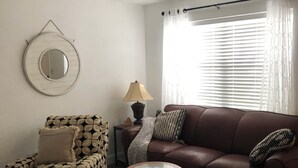 A living room with a brown leather sofa, a patterned armchair, a round coffee table, a side table with a lamp, a wall-mounted mirror, and a large window with blinds.