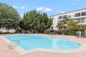 A swimming pool with a metal ladder, surrounded by a patio area with chairs and a building in the background.