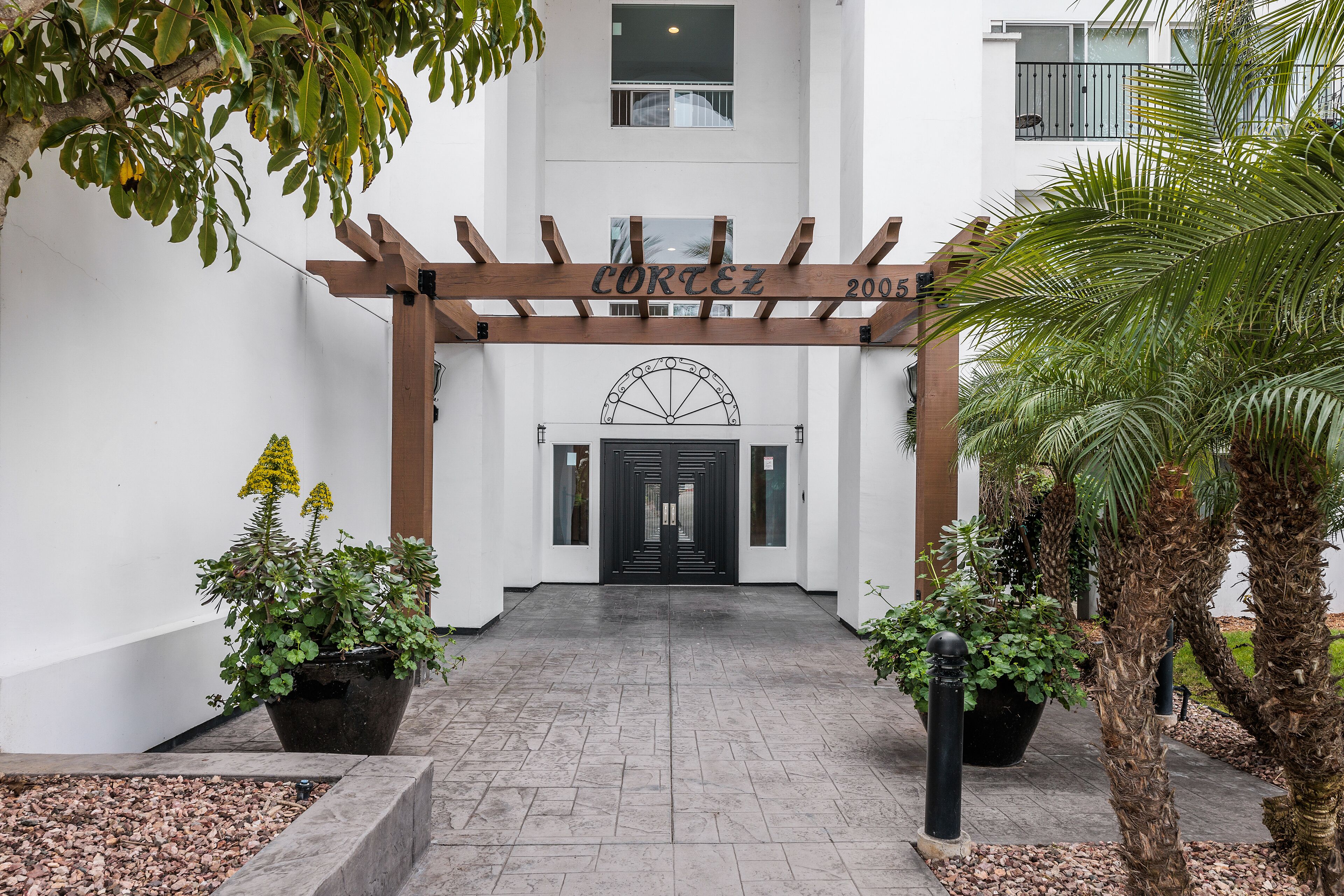 Ein Gebäudeeingang mit einer hölzernen Pergola, die über der Tür verläuft, einem schwarzen Tor und Kübelpflanzen auf beiden Seiten.