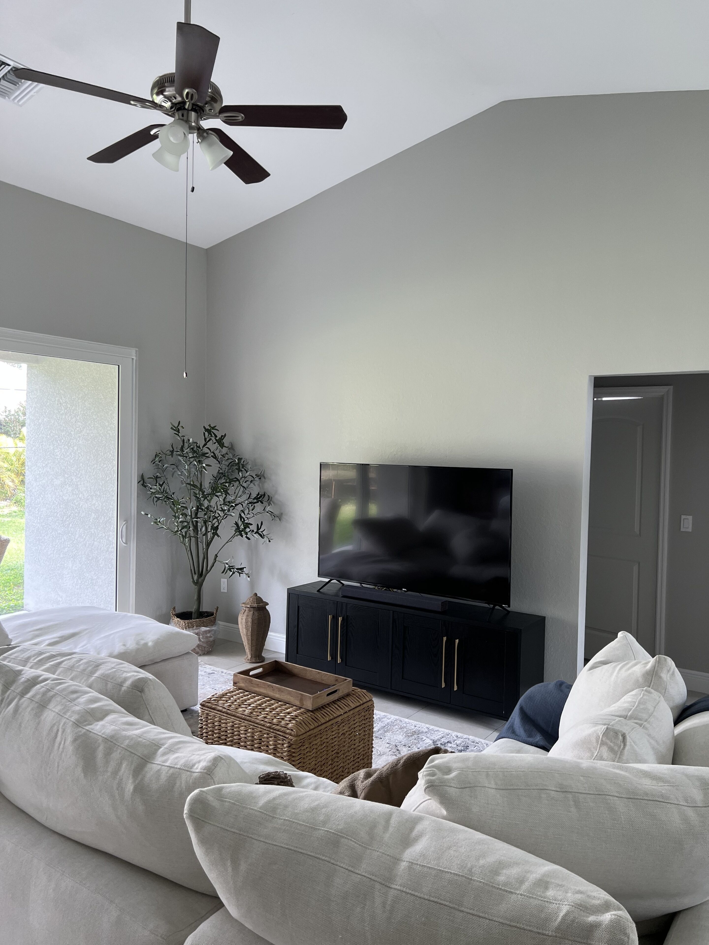 A modern living room with a large sectional sofa, a flat-screen TV on a black cabinet, a ceiling fan, and a potted plant.