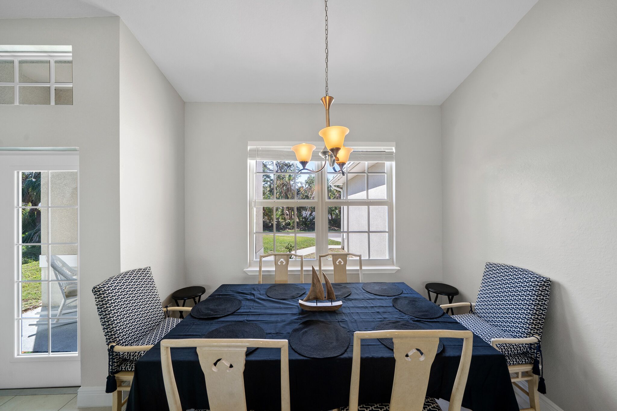 A dining room with a table set for six, a chandelier, and a view of the outdoors through large windows.