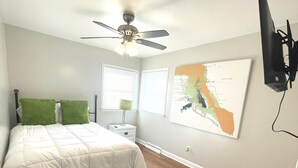 Quarto com cama, ventilador de teto, mesa de cabeceira, abajur e televisão fixada na parede.