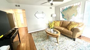 Uma sala de estar com uma televisão de tela plana, um ventilador de teto, um sofá, uma mesa de centro com um vaso de flores e um grande quadro abstrato na parede.