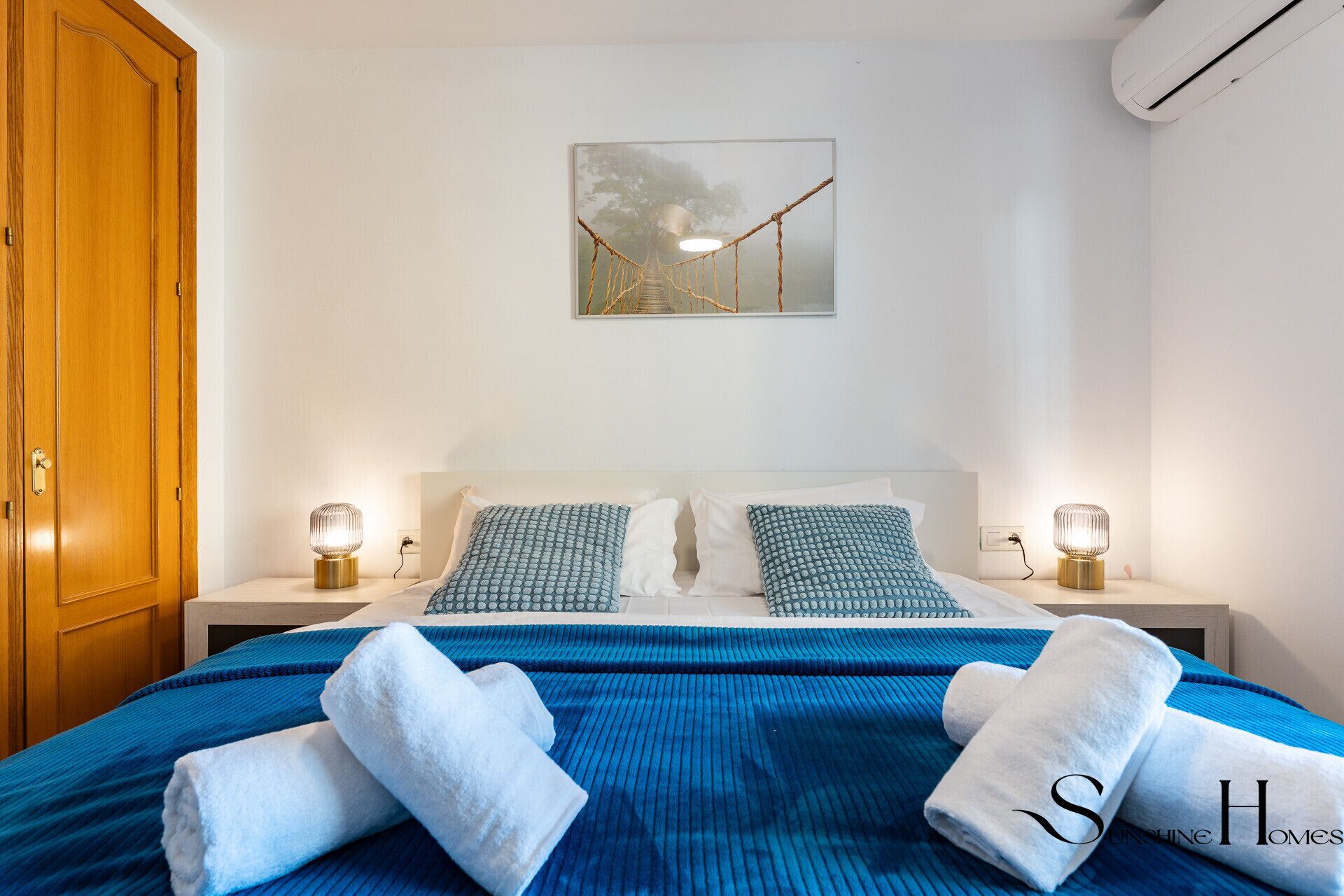 A bedroom with a bed, two bedside tables, a wall-mounted artwork, and an air conditioning unit.