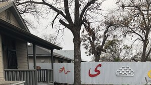 A building with a covered porch, a fenced area with a logo, and a gravel driveway. - Alamo City Former Library Historic Home, Parking, BBQ, Firepit, Gated Side-Yards (San Antonio)