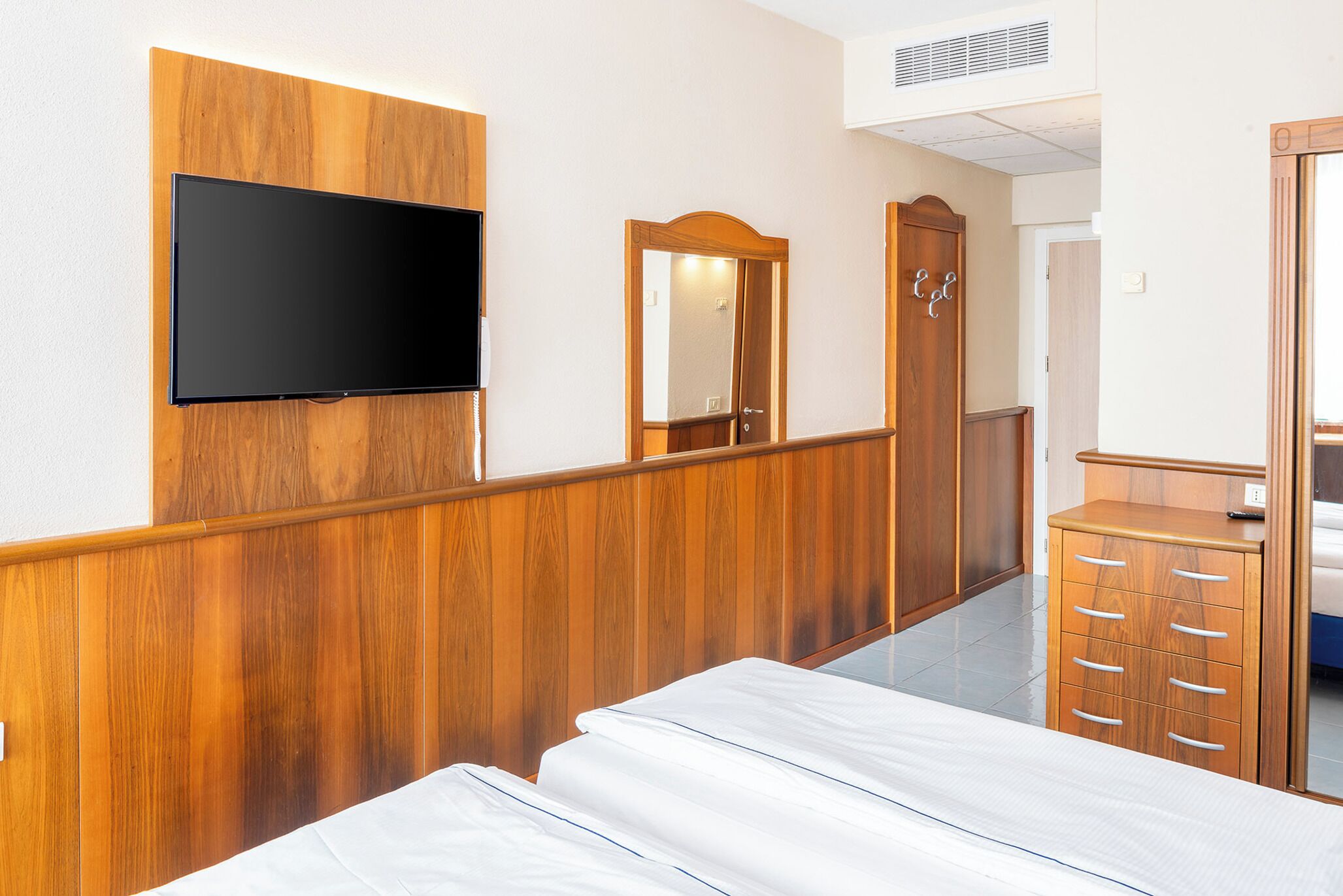 A hotel room with a wooden wall panel, a flat-screen TV mounted above a bed, a wooden dresser with drawers, and a mirror.
