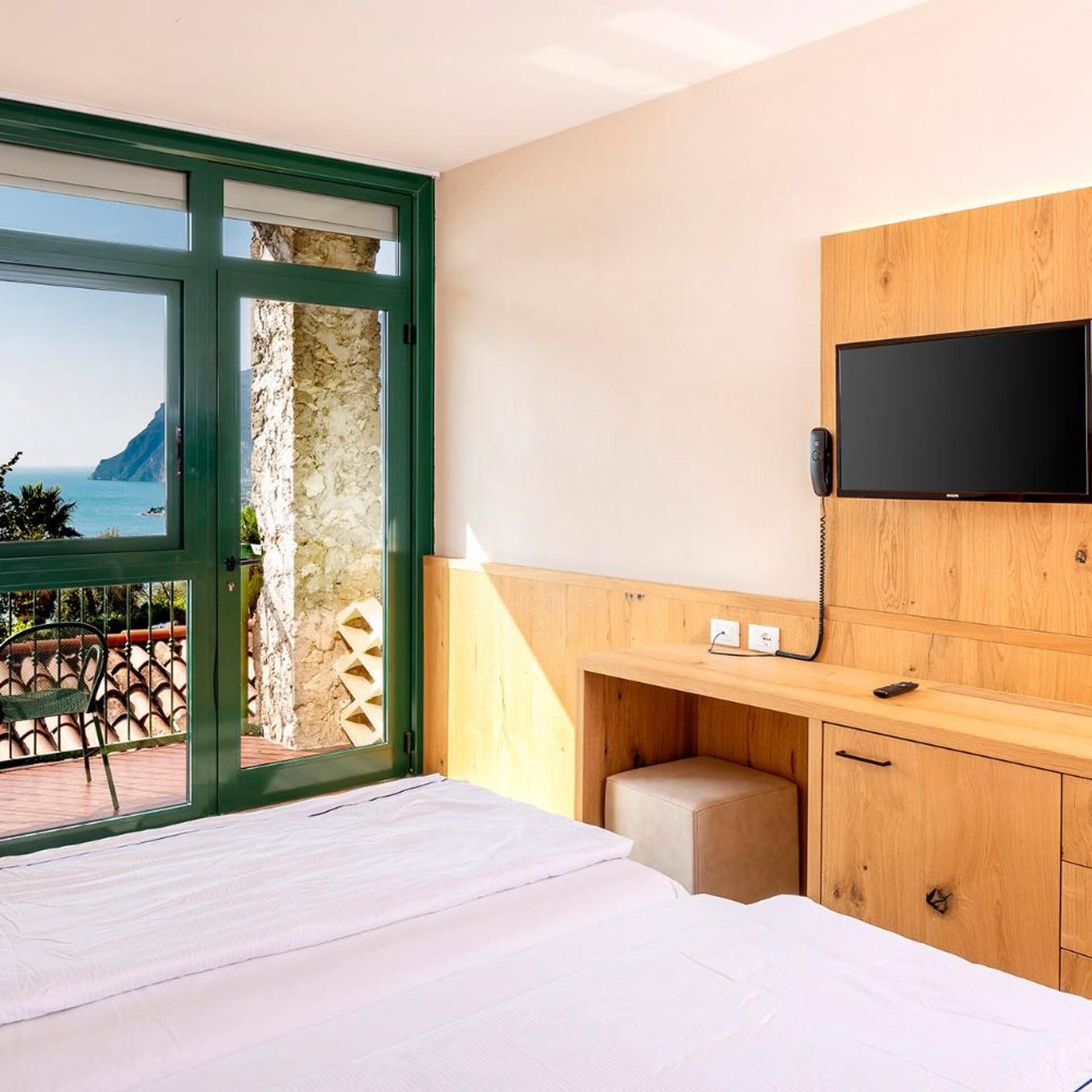 A bedroom with a bed, a TV, a wooden desk, and a view of the sea.