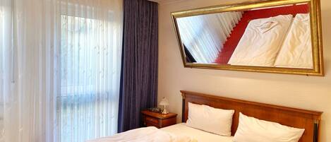Habitación de hotel con cama doble, mesita de noche, espejo y ventana con cortinas traslúcidas.