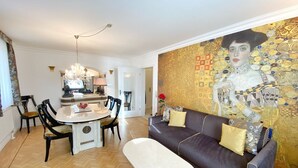 Un coin repas avec une table ronde, des chaises et un lustre. On y trouve également un canapé avec des coussins et une grande fresque murale décorative.