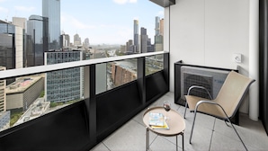 A balcony with a city view, a chair, a small table with a glass of wine and a book, and a modern building in the background.