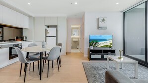 A modern living room with a flat-screen TV, a dining area with a table and chairs, a kitchen with white cabinets and appliances, and a large window with a view of the beach.
