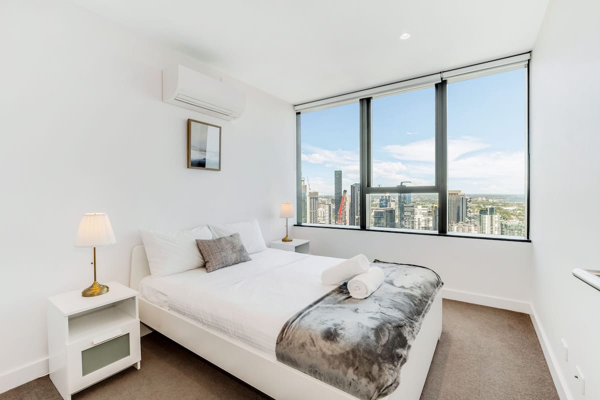 Quarto moderno com cama grande, mesa de cabeceira, abajur, janela com vista para a cidade e ar-condicionado instalado.