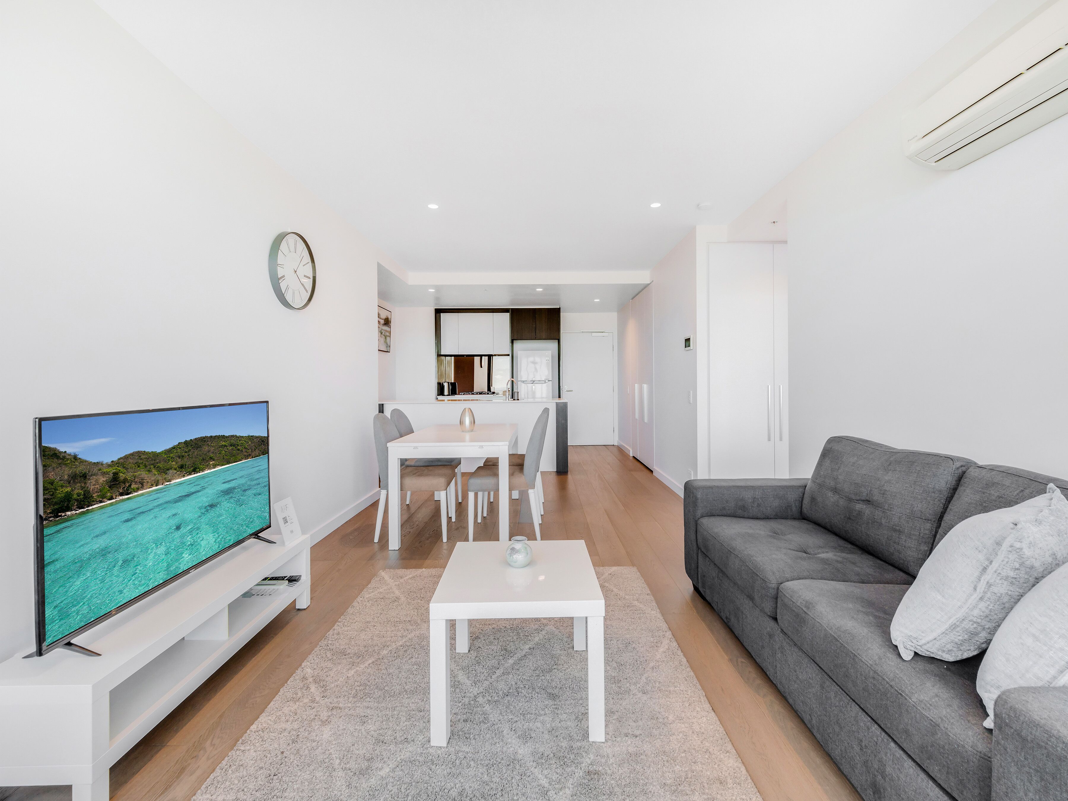 A modern living room with a flat-screen TV, a gray sofa, and a dining area with a white table and chairs.