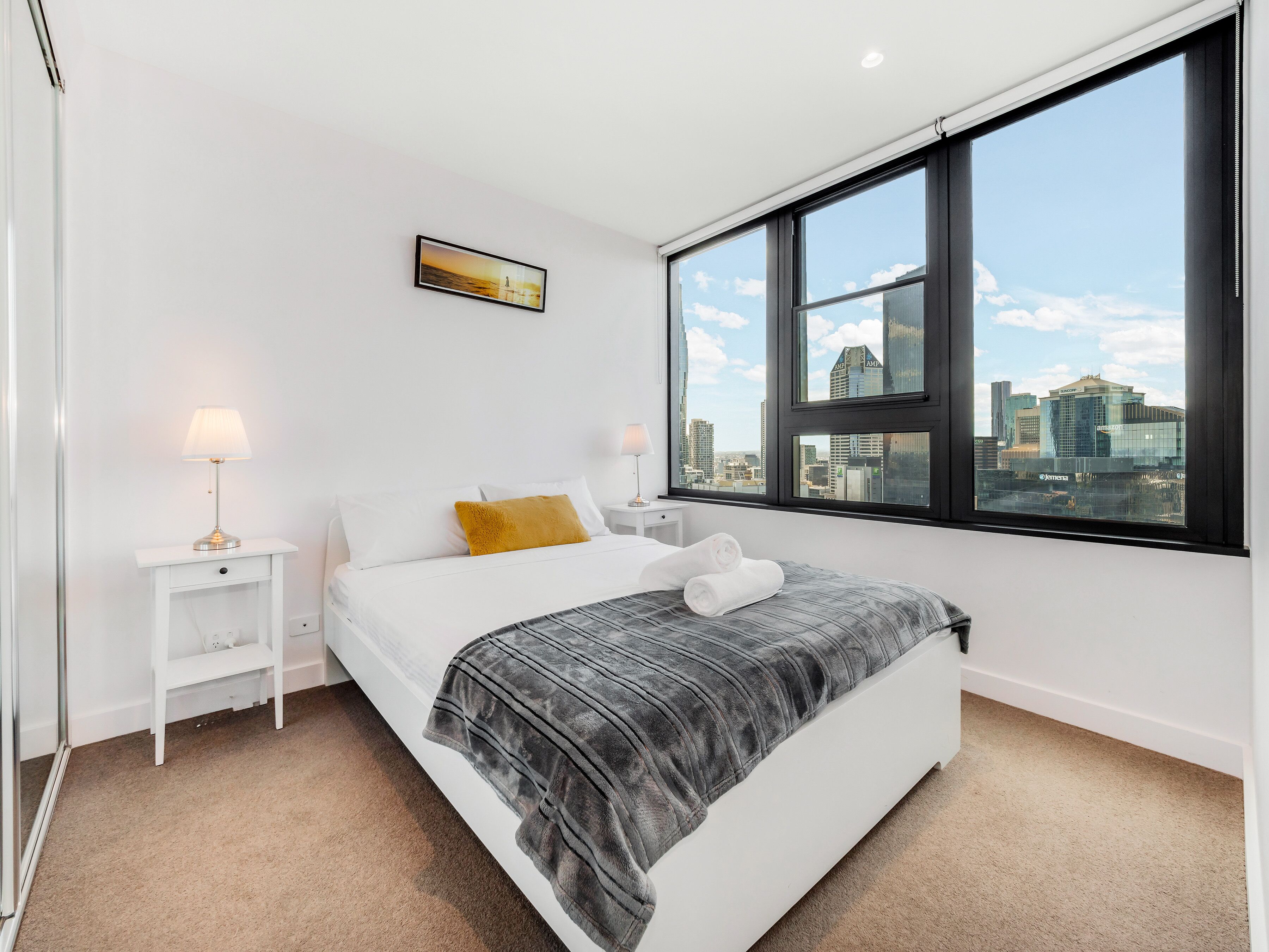 A modern bedroom with a large window, a bed with white bedding and a gray blanket, a nightstand with a lamp, and a cityscape view.