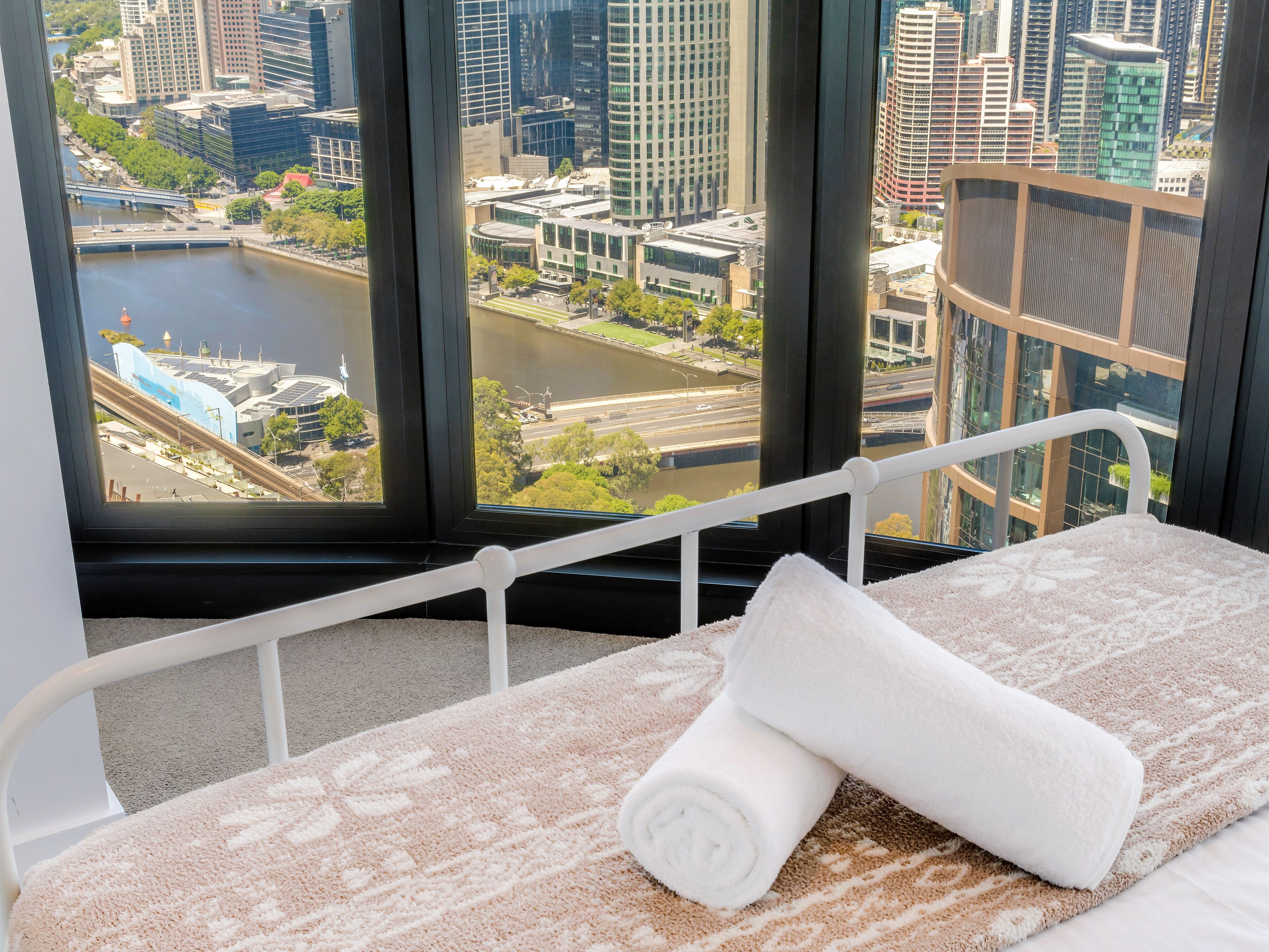 Una cama con una toalla enrollada, vistas de la ciudad a través de amplias ventanas y una barandilla de balcón.