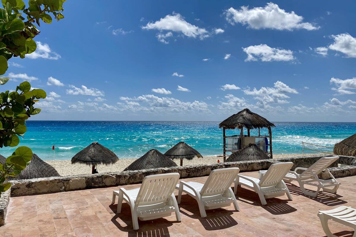 Una playa con sillas de playa blancas y sombrillas de paja, con vista al mar azul y transparente.