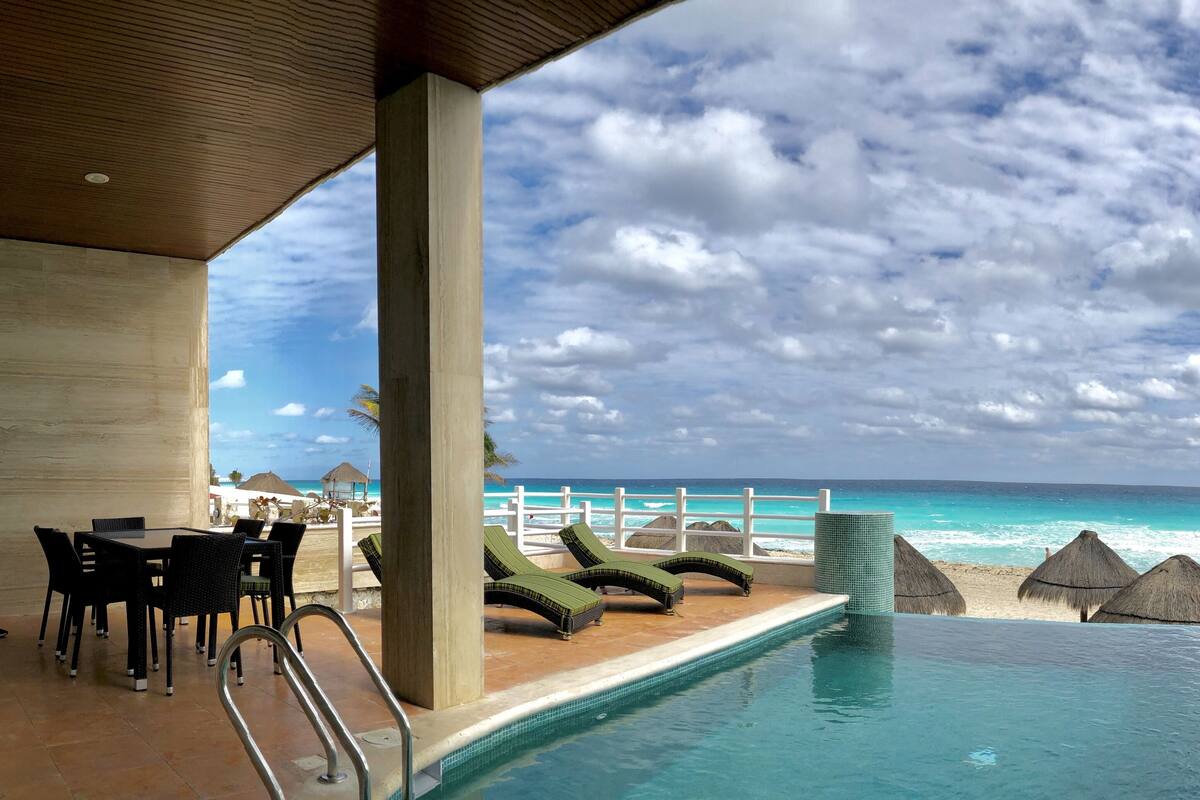 Zona de piscina con sillas de descanso y una mesa, con vistas a una playa de aguas turquesas y cielo nublado.