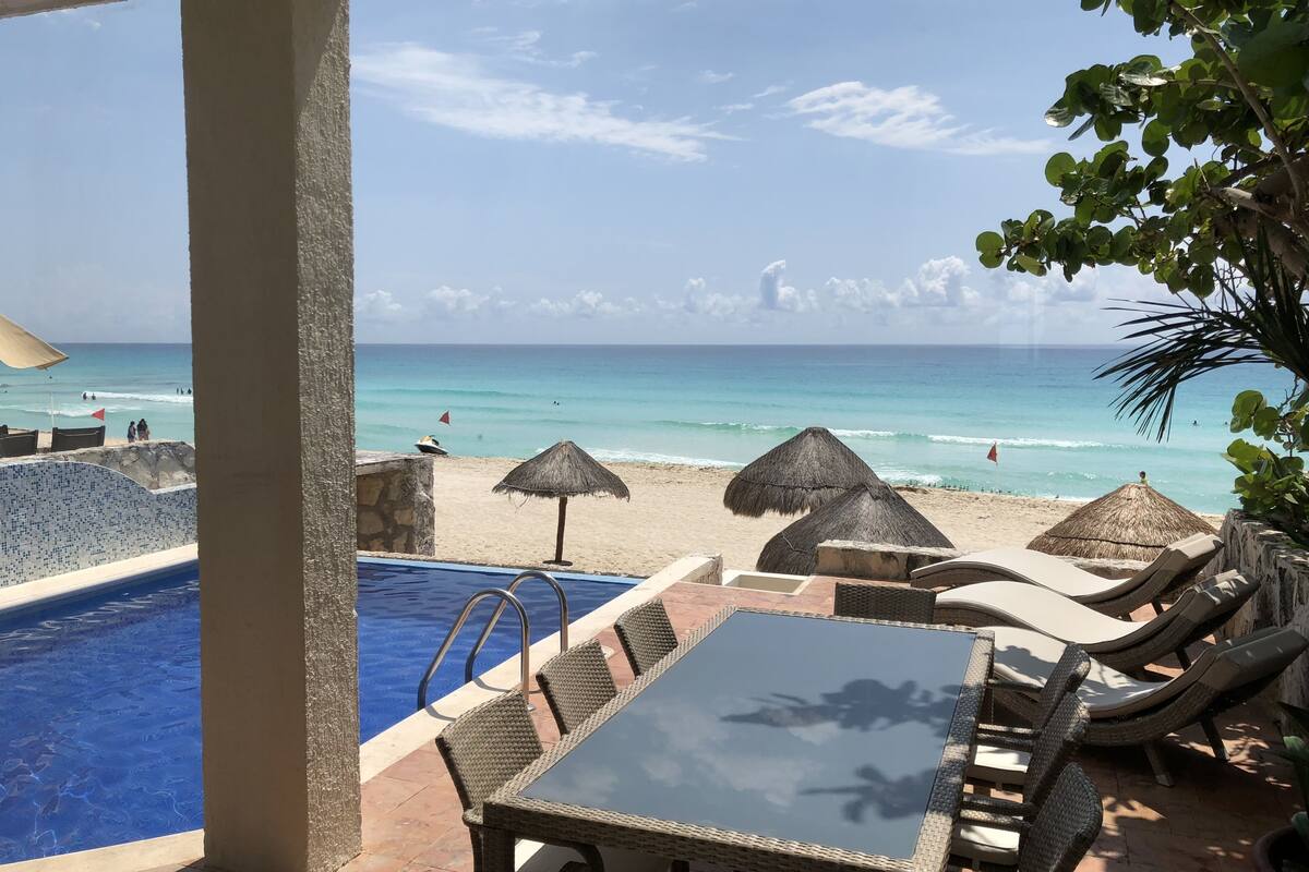 Zona junto a la piscina con mesa y sillas de vidrio, vista a la playa y sombrillas de paja.