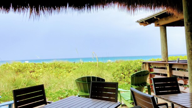 Ein Sitzbereich im Freien mit Tischen und Stühlen mit Blick auf einen Strand, viel Grün und das Meer.