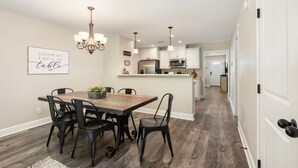 A dining area with a wooden table and black chairs, a chandelier, and a kitchen in the background.