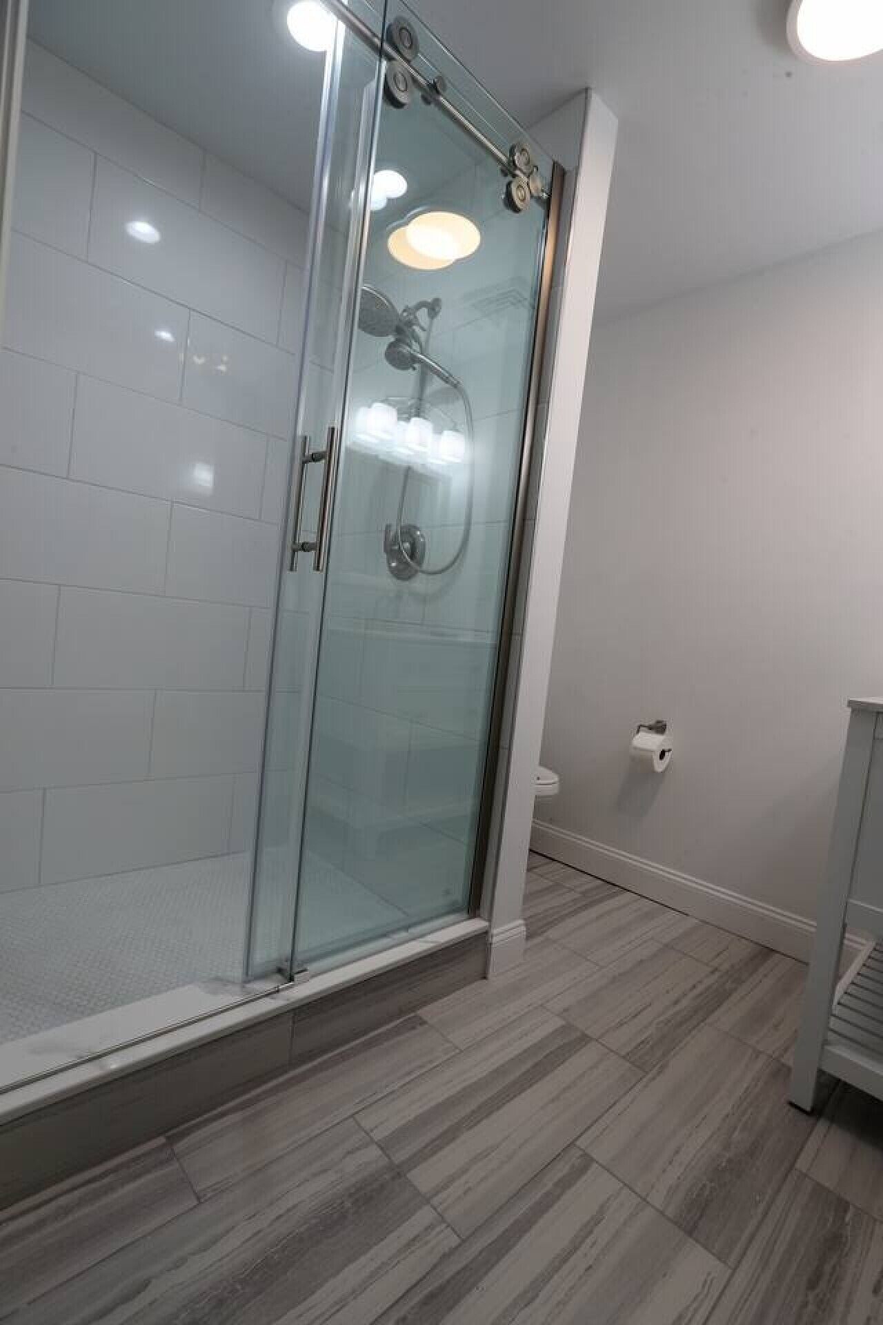 Une salle de bain moderne avec une douche à parois de verre, des murs recouverts de carreaux blancs et un plancher en bois.