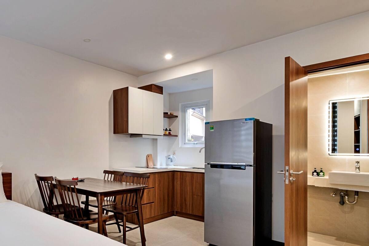 Una habitación de hotel con una cama, una mesa de comedor con sillas, una zona de cocina con nevera y lavabo, y un baño con espejo y lavabo.