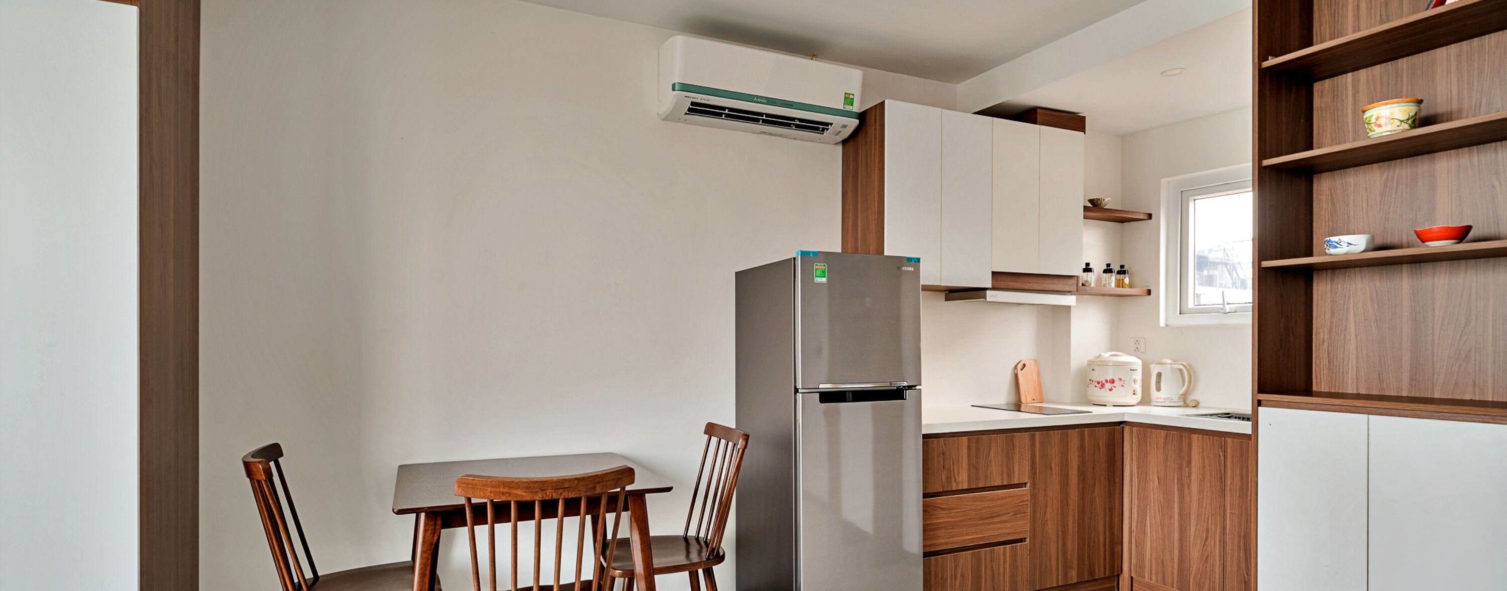 A compact kitchen with a refrigerator, wooden cabinets, a dining table with chairs, and a window with shelves above.