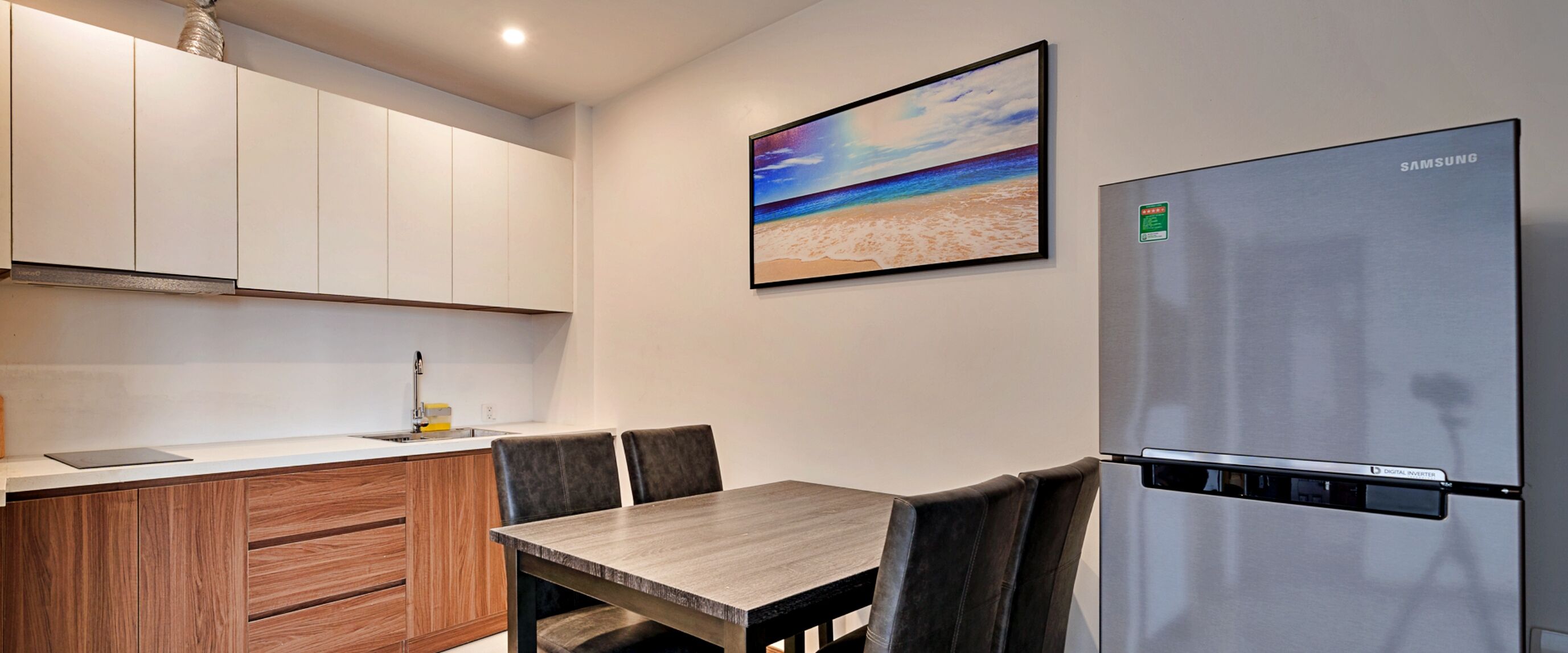 A modern kitchen with white cabinets, a stainless steel refrigerator, a dining table with chairs, and a beach scene painting on the wall.