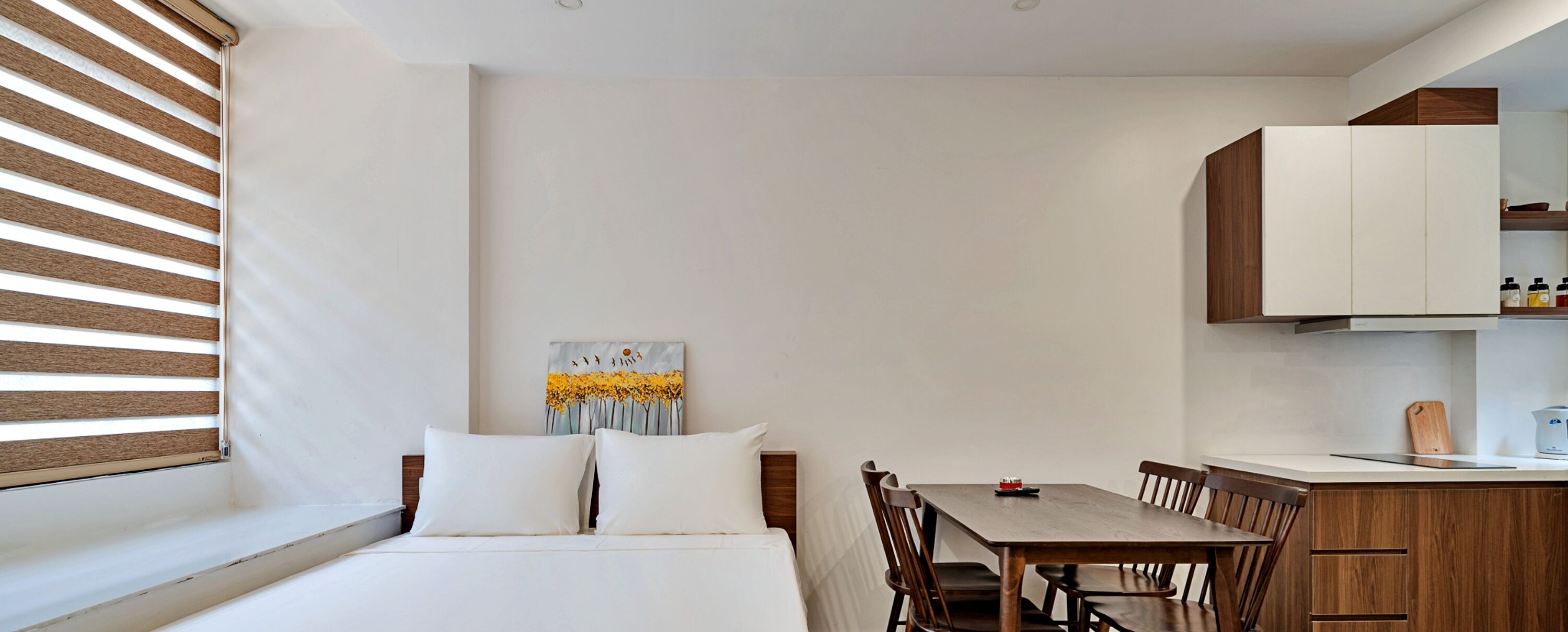 A bedroom with a bed, a dining table, and a kitchen area with cabinets.