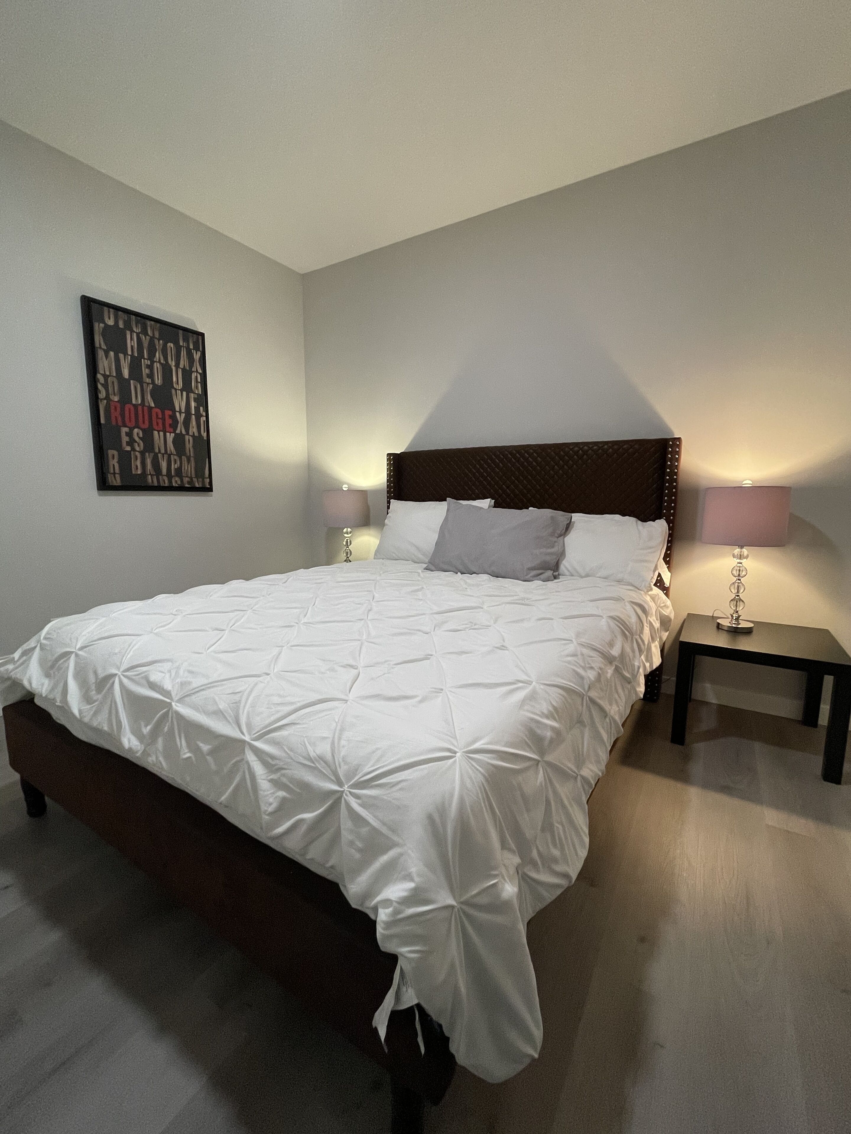 Een net opgemaakt bed met een wit gebogen dekbed, een houten hoofdbord en twee nachtkastjes met lampen.